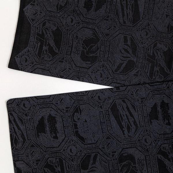A detail of a charcoal black scarf with a grid of octagonal vignettes also printed in black. Each features an abstract composition of a car, hot air balloon, or another scene. Two ends of the scarf are folded over one another, slightly overlapping.