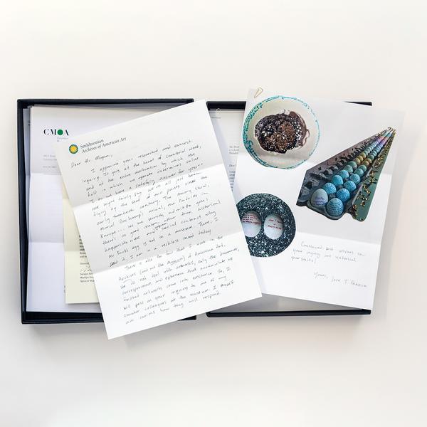 A black box sits open facing up against a white background. Stack of shuffled letters lie within the box's enclosure. A hand-written letter on Smithsonian Archives of American Art letterhead lies overtop with another document showing blue eggs in a seemingly endless carton near two circles with mineral-like textures.