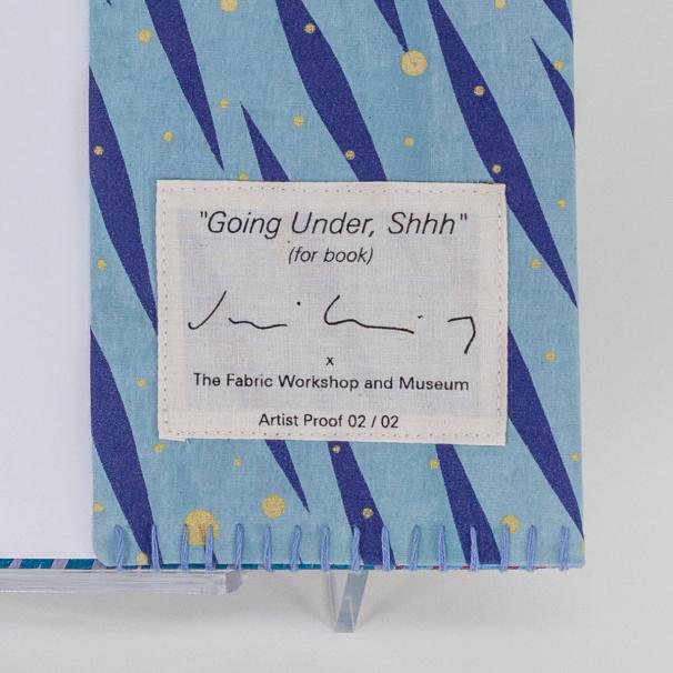 A photo of a hardback book open to the first blank pages featuring the slip of the fabric book cover with a hand-stitched seam and a vibrant pattern of diagonal dark blue brush-like streaks and scattered yellow dots on a light blue background. There is a cream fabric label that has been screenprinted to feature the title of the edition, the artist's signature, and the artist proof number