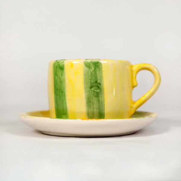 A small handmade teacup with vertical stripes in yellow and green sits on top of a saucer against a white background. The mug has a yellow handle. The saucer has color on the inner face and is white on the underside.