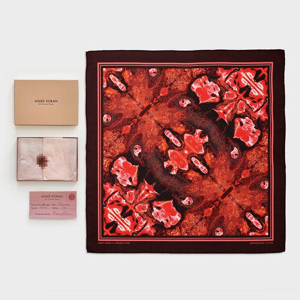 An arrangement of objects against a white ground. On the right: a square scarf with an abstract red pattern that is mirrored along the diagonal corners and bordered with thick maroon and thin pink stripes; on the left from top to bottom: a rectangular beige box lid that reads "Aimee Koran for Corner Store," the interior of the box with tissue paper adhered with a pixelated nipple sticker; and a dark pink certificate of authenticity.