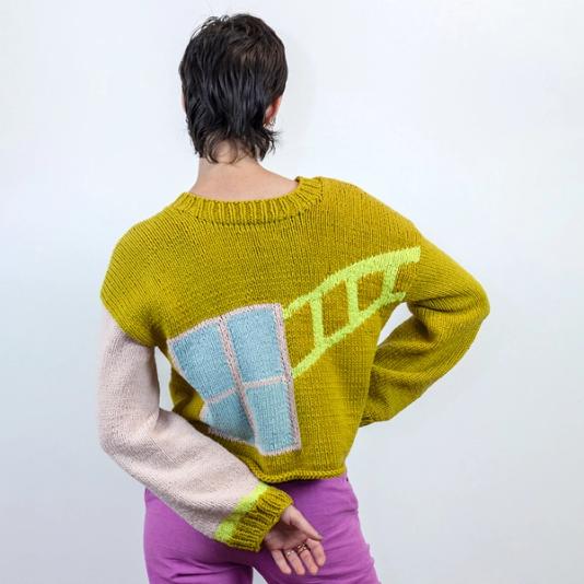 A person with dark short hair wears a long sleeve sweater with her right elbow out and her left arm folded behind her. The body of the sweater is a medium toned yellow green. The left sleeve is pink with a green cuff and a neon yellow accent next to the cuff. On the back of the sweater, there is the light pink implication of a window with a ladder of neon yellow light emanating toward the shoulder.