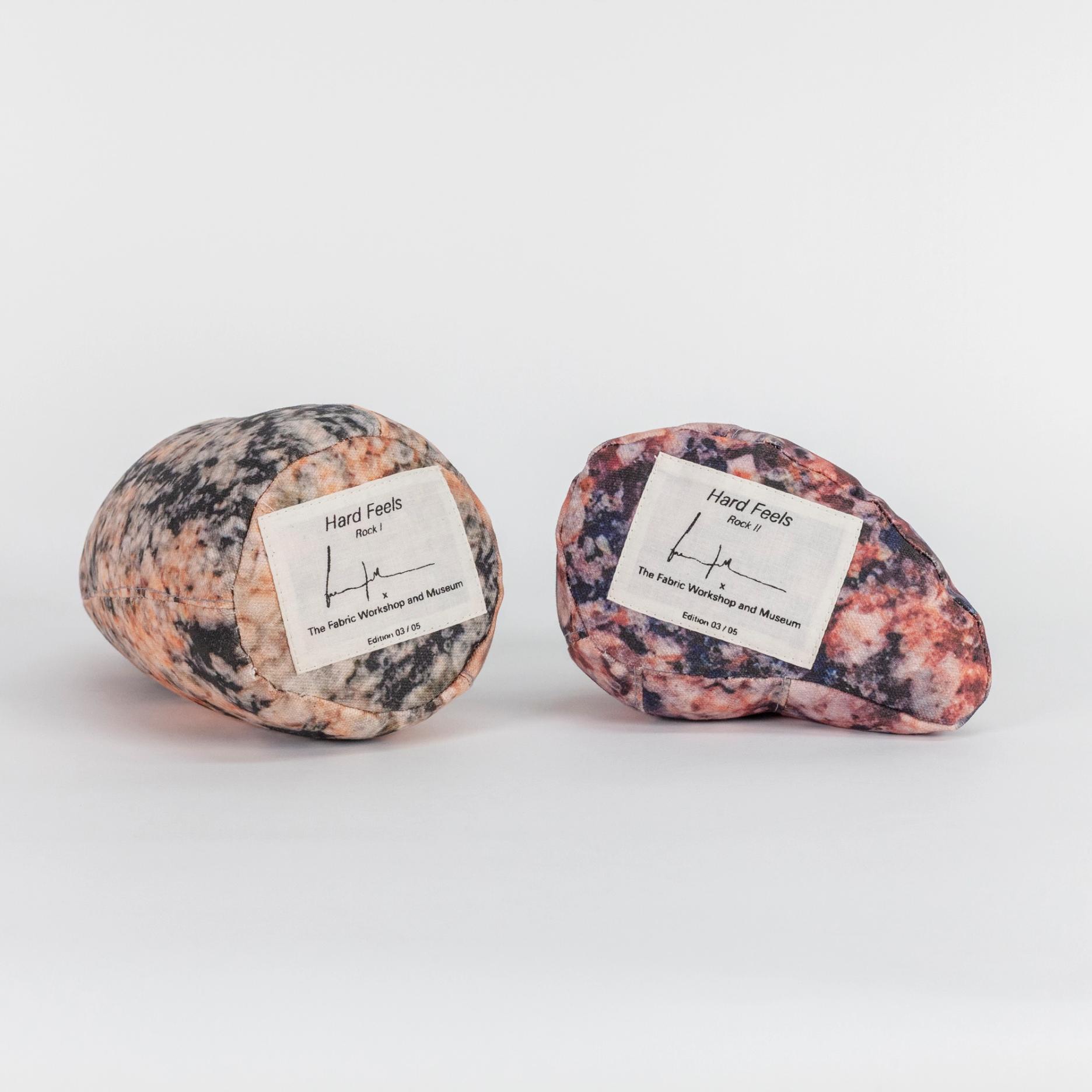 A photograph of the bottom sides of two small soft sculptures covered with patterns that resemble polished stone of different hues. Each one has a label reading the title of the edition (Hard Feels), the artist's signature, and edition number.
