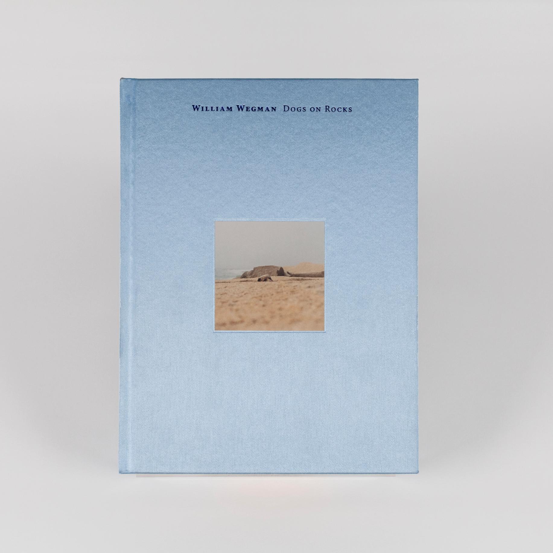 A sky-blue book, taller than it is wide, featuring a square photograph of a beach at its center. The text reads, "William Wegman, Dogs on Rocks."