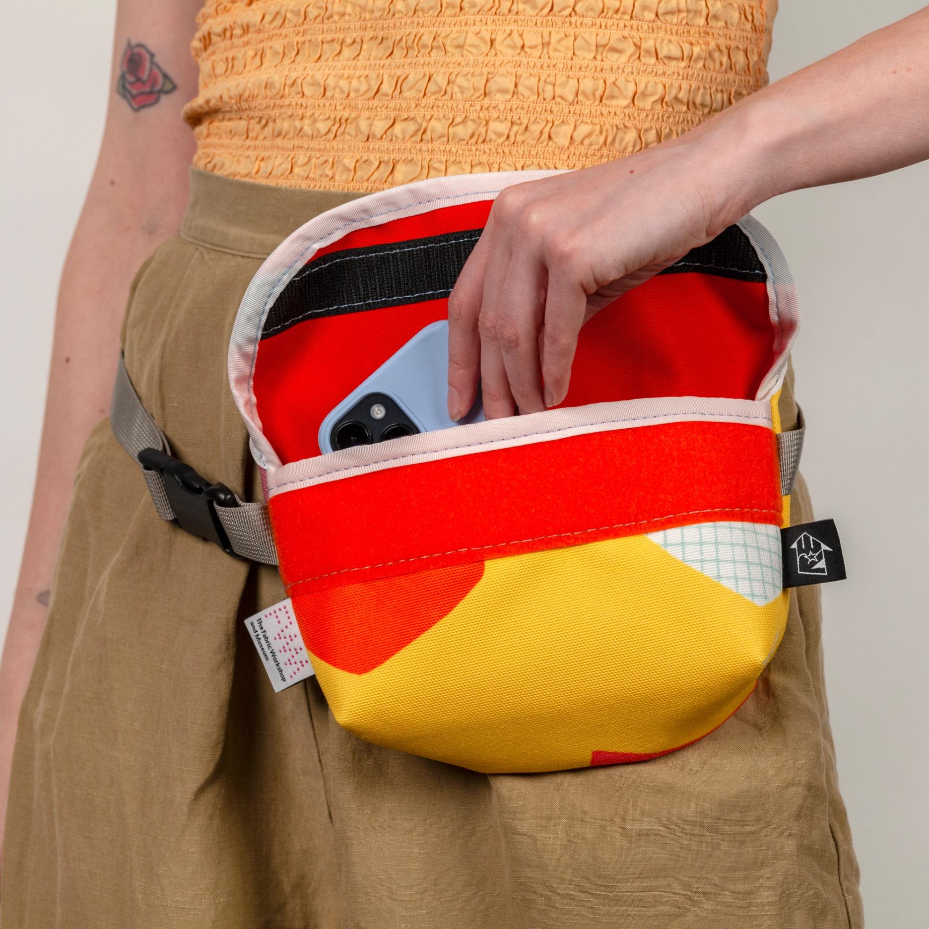 A model wearing a shallow, wide, vibrant yellow bag at her hip (like a fanny pack) with bold abstract shapes (bright orange and white with a light blue grid pattern). The model is holding open the top flap which has a bright orange lining and black velcro closure with a white seam along the flap and opening of the bag. The bag has a shiny gray strap and plastic black buckle.