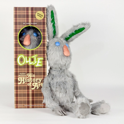 A stuffed plush gray rabbit sits upright against a plaid collector's box. The rabbit has round blue eyes, an oblong peach nose and long green shapes on the interior of its ears. The brown plaid box reads "Ollie" and "Jayson Musson's His History of Art" and has a circular viewing window that reveals another rabbit puppet inside.