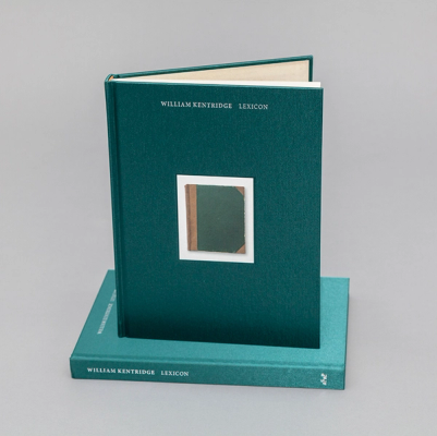 A photograph of a green book standing upright, its pages slightly opened, atop another copy of the same book. The cover features a smaller photo of an aged green book with brown binding. The title at the top reads in an all-caps serif font, "William Kentridge Lexicon" 