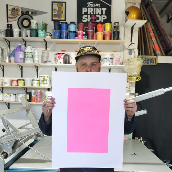 A portrait of John Ream, a white man who is wearing a black cap and holding a print featuring a pink rectangle, standing in a print studio. Jars of ink stack up on shelves behind him.