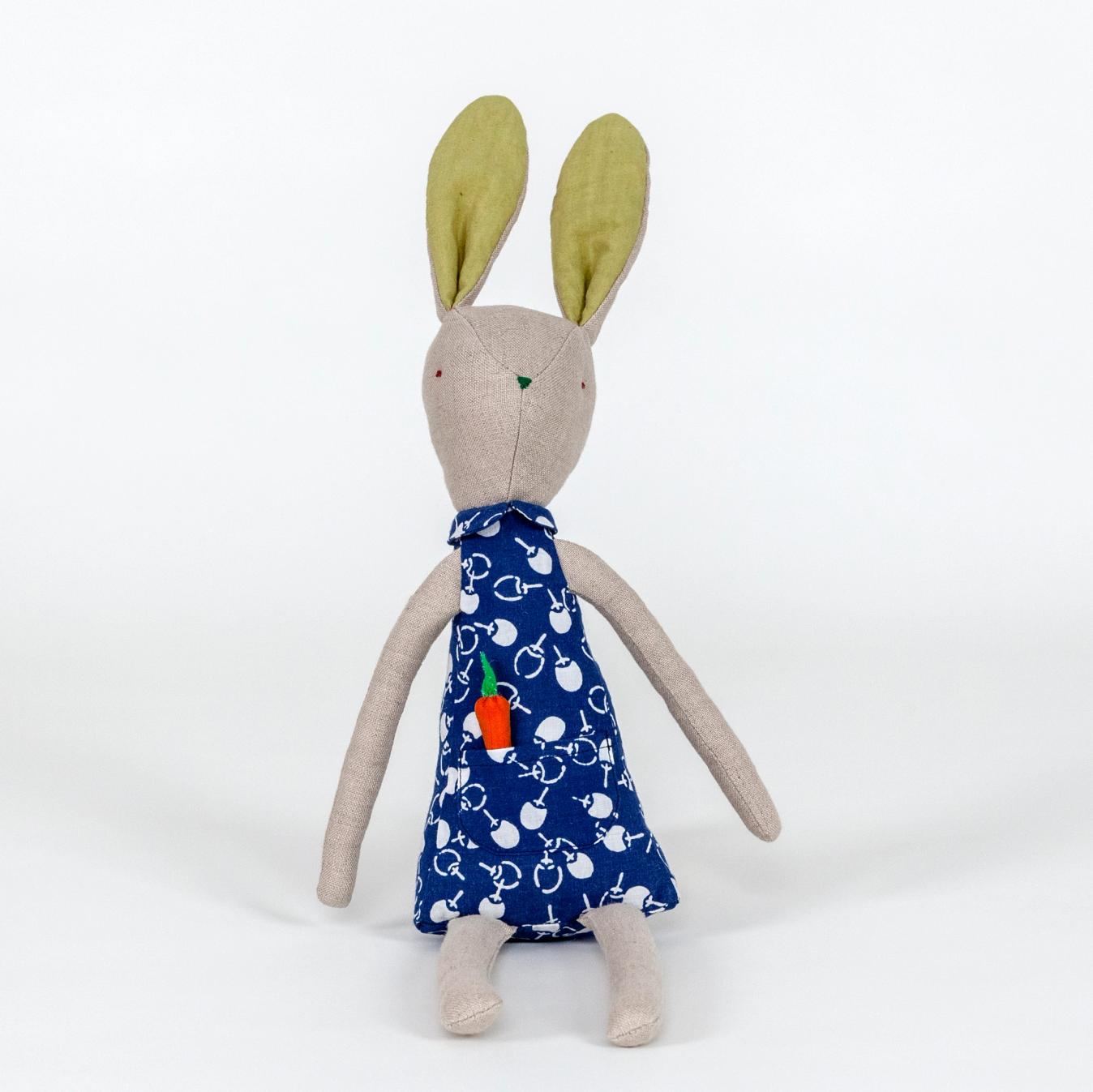 A photograph of a plush rabbit made of light beige woven fabric characterized to have a pointy nose, triangular shaped body, and long thin arms and legs. The body of the rabbit is made of a primarily blue patterned fabric and includes a pocket with a removable plush orange carrot.