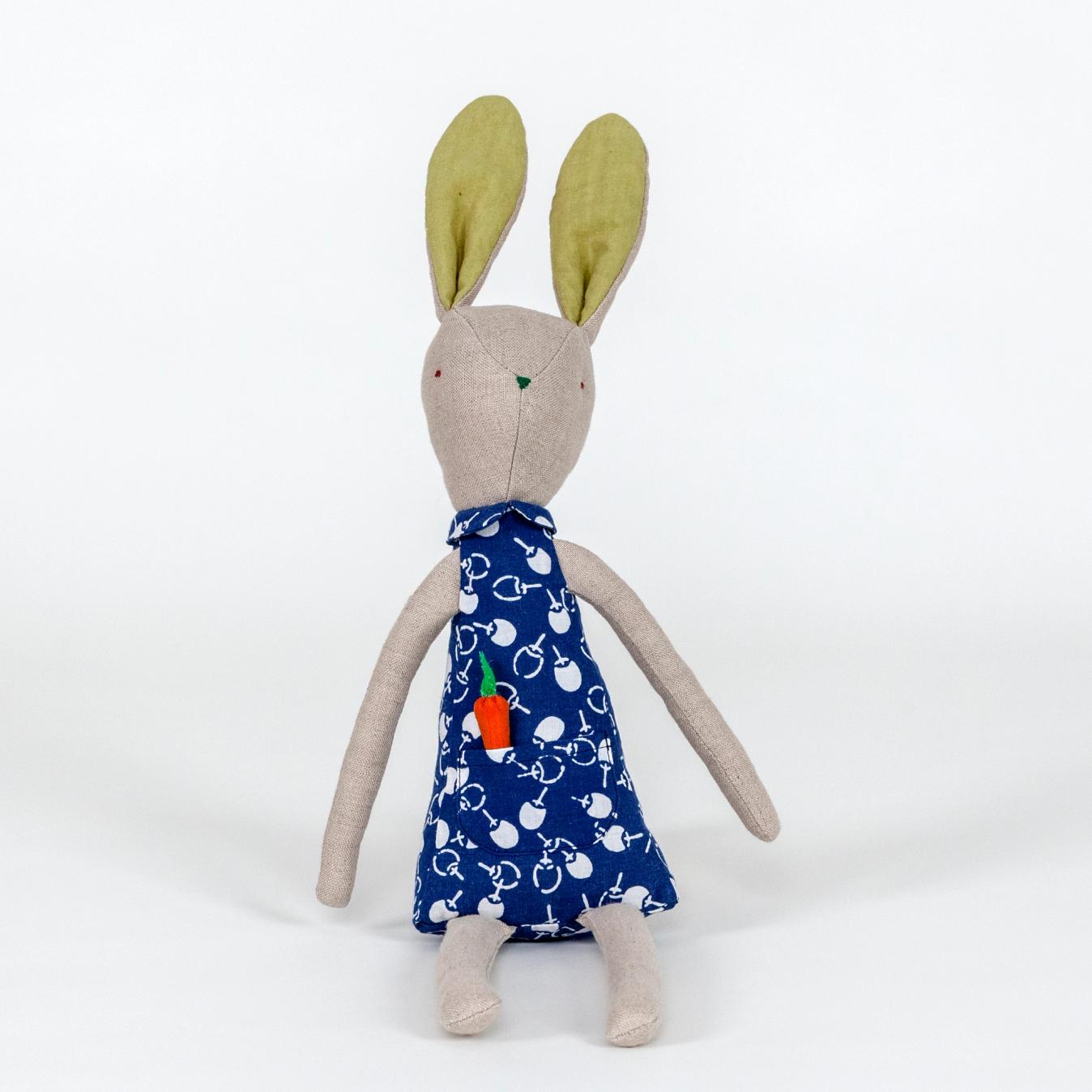 A photograph of a plush rabbit made of light beige woven fabric characterized to have a pointy nose, triangular shaped body, and long thin arms and legs. The body of the rabbit is made of a primarily blue patterned fabric and includes a pocket with a removable plush orange carrot.