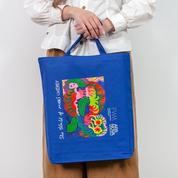 A model holds a blue tote in front of her, with one hand holding the straps and the other resting overtop. The tote features a colorful abstract painting composed of organic shapes. It reads "The World of Moki Cherry" and features the logos of The Fabric Workshop and Museum and Ars Nova Workshop.