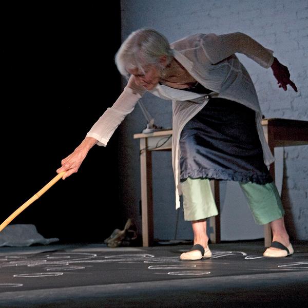 A photograph of an older white woman leaning over to draw or point with a long stick. She is wearing a thin white sweater buttoned at her waist over a dark skirt and light green pants.