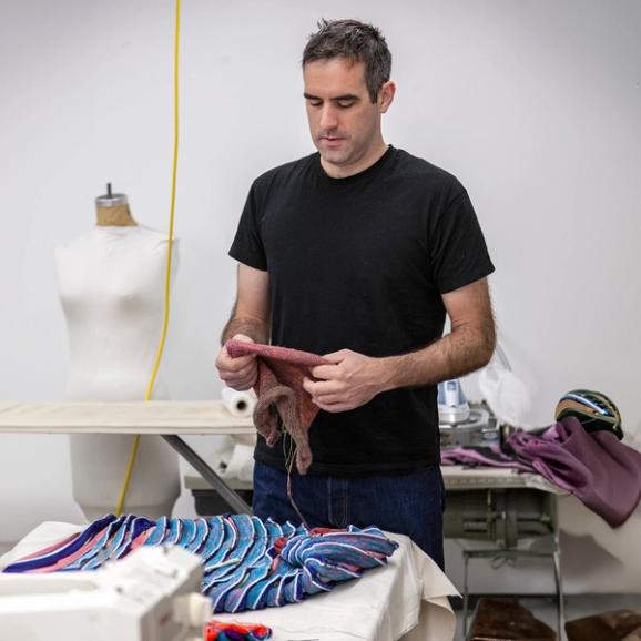 A photograph of a white man with short dark hair and wearing a black t-shirt standing over a table in a studio. He is holding a woven fabric material. On the table is a colorful pleated fabric of blues, purples, and reds. Behind him to his right is a mannequin torso.