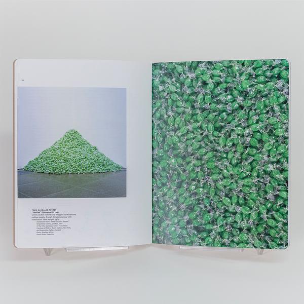 A photograph of a spread of an open book. On the left there is a photograph of a big pile of green individually wrapped candies on the floor and against a white wall. Below the photo is a description of the work. On the right there is a close up photo of the individually wrapped candies densely packed and taking up the whole page.