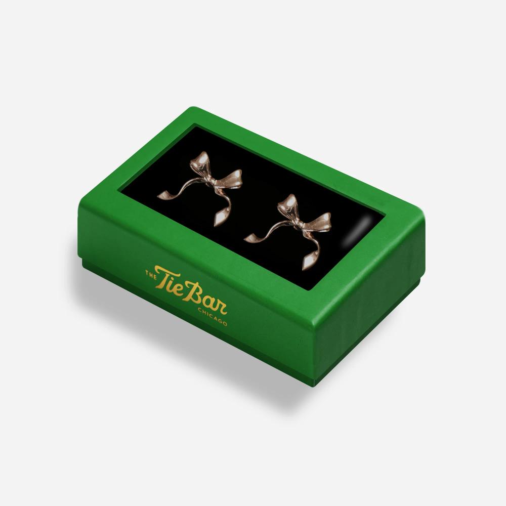 A rectangular box with a green border features a pair of rose gold colored cuff links with a decorative bow inside.