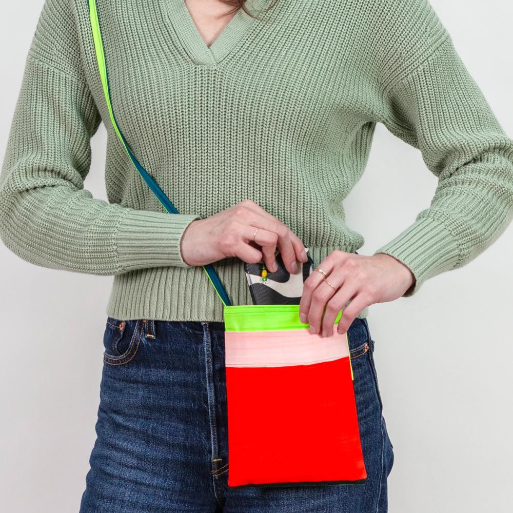 A model carrying a small, rectangular crossbody bag at the hip that features bold light pink, red, and neon chartreuse stripes. The thin strap is neon chartreuse and teal. The model is pulling a smartphone out of the bag.