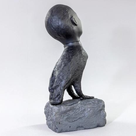The back of a small sculpture of a creature composed of a human head and a bird's body perched on a small rock.