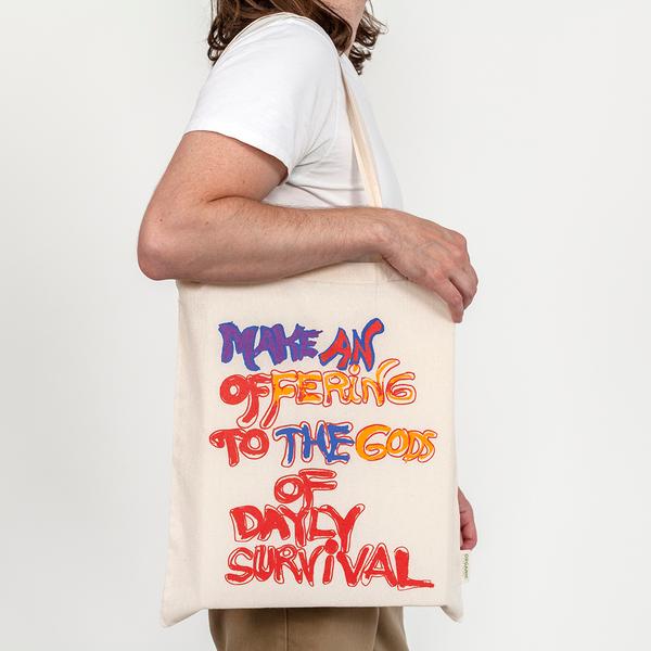 A model carries a cream-colored tote on his shoulder that reads in a hand-drawn message, "Make An Offering to the Gods of Daily Survival." The word daily is spelled d-a-y-l-y. Half the words appear in red while others are colored in with purple, pink, burnt yellow, and blue.