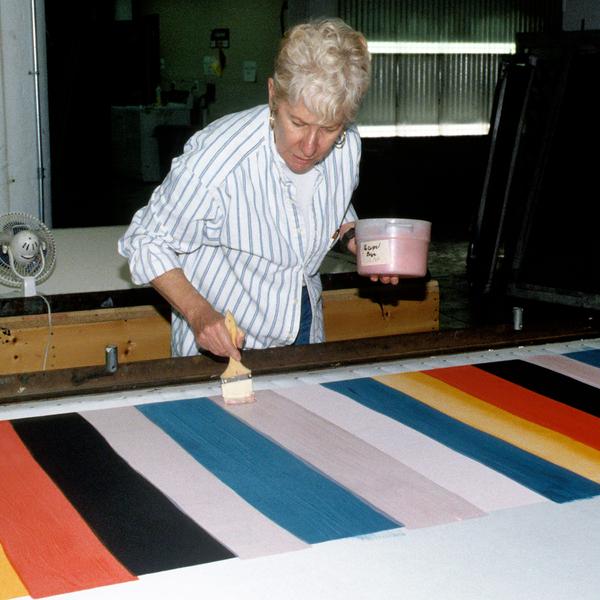 An photograph taken with flash spotlighting on Mary Heilmann, an older white woman with very light blonde hair standing over a table while holding a tub of ink in her left hand and a brush in her right hand. She is brushing stripes on a multi-colored striped pattern featuring blue, yellow, red, black, white and light pink. Some of the stripes may not have been painted yet.