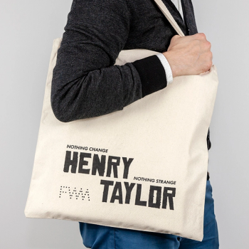 An image of someone facing to the right and holding the tote bag on their shoulder. They are wearing a grey long sleeved shirt and are holding the strap of the bag in their hand. The tote bag has the back side facing outwards, and the screenprinting in black says "Nothing Change, Nothing Strange" alongside the artist's name "Henry Taylor" in large font at the bottom of the tote. In the bottom left corner, is The Fabric Workshop and Museum's logo, "FWM" written in dots.