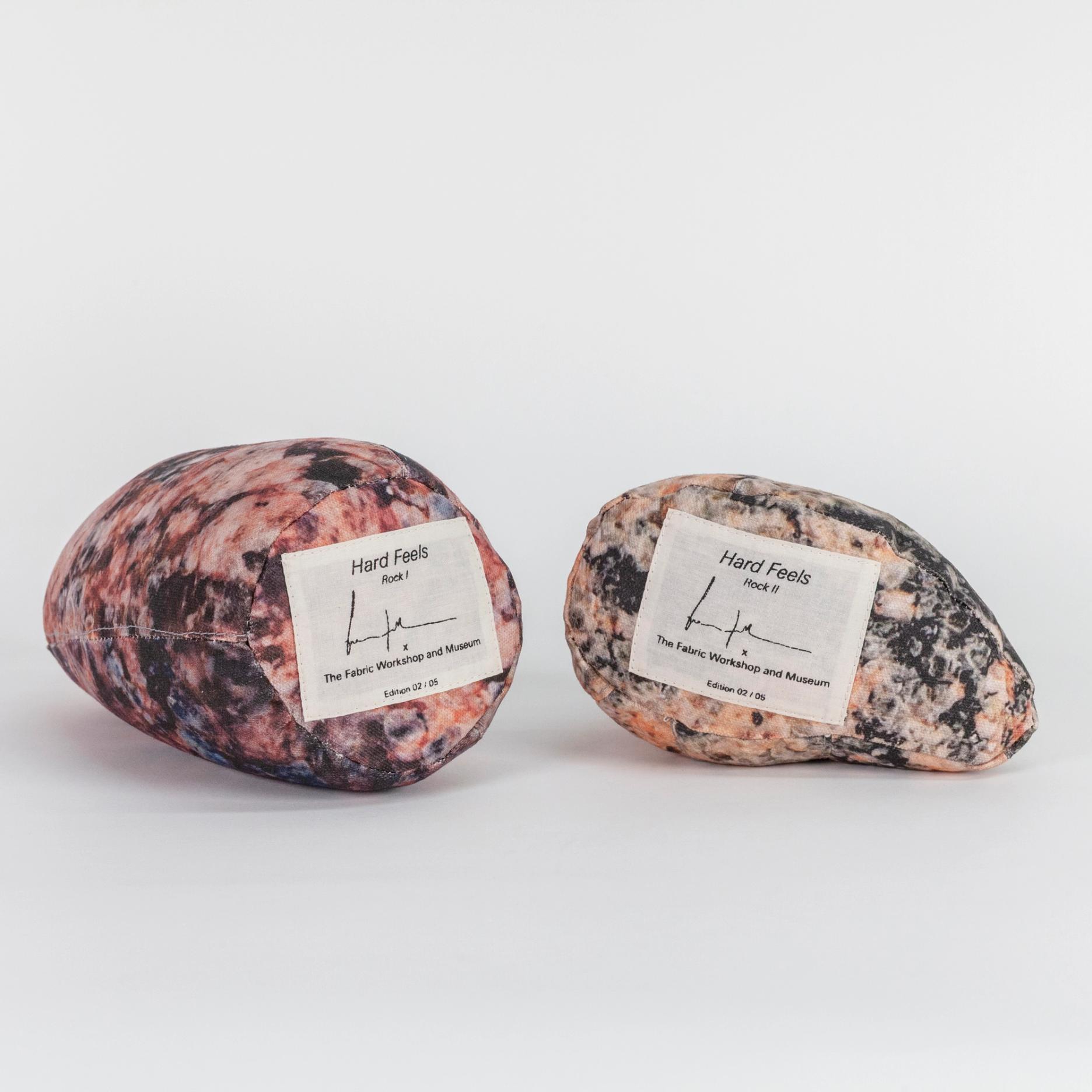A photograph of the bottom sides of two small soft sculptures covered with patterns that resemble polished stone of different hues. Each one has a label reading the title of the edition (Hard Feels), the artist's signature, and edition number.