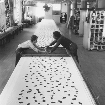 A black and white photograph of two people—a woman on the left and a man on the right—screenprinting a pattern of small black shapes from each side of a long printmaking table. 