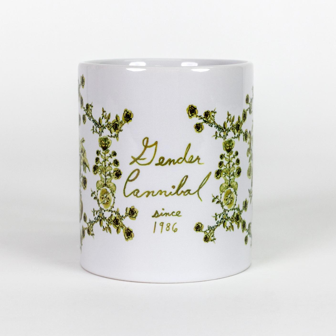 A white ceramic mug with "Gender Cannibal since 1986" written in green, watercolor-like script. Surrounding the text is a hand-illustrated green toile-like pattern made up mostly of roses but also features twin fetuses and rabbits.