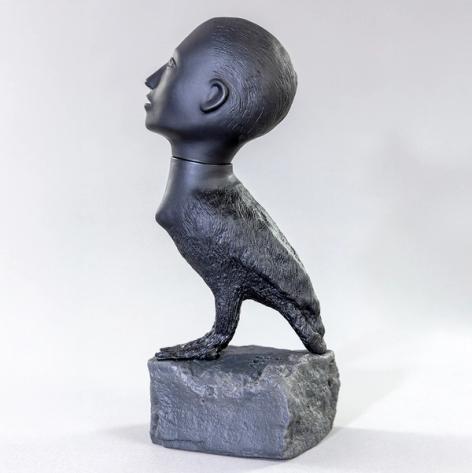 A small sculpture of a creature composed of a human head and a bird's body perched on a small rock.