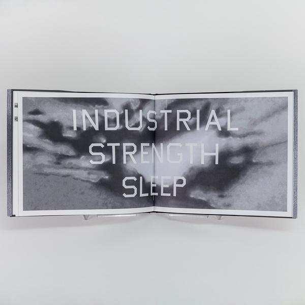 A photograph of an open book showing a white and black stylized cloudy sky with overlayed white text reading "INDUSTRIAL STRENGTH SLEEP"