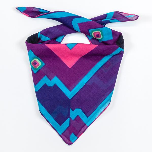 A square scarf folded over with ends ready to be tied around one's neck. The scarf is mostly purple, with varying shades of blue and a bright pink border around the designs in the center.