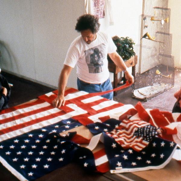A man standing over a table with multiple American flags. He is weaving together the red and white stripes of two of the American flags.