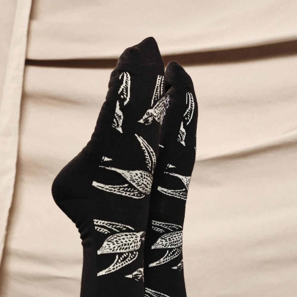 A detail of a person modeling a pair of black knit dress socks with a pattern of white illustrated Shomoro birds flying in different directions.