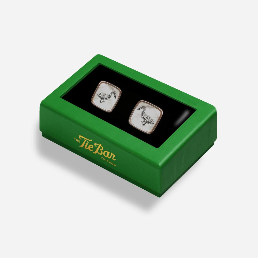 A rectangular box with a green border features a pair of square white pearlescent cufflinks with a rose gold border and clips. On the pearlescent face is a black line illustration of a Bata bird.