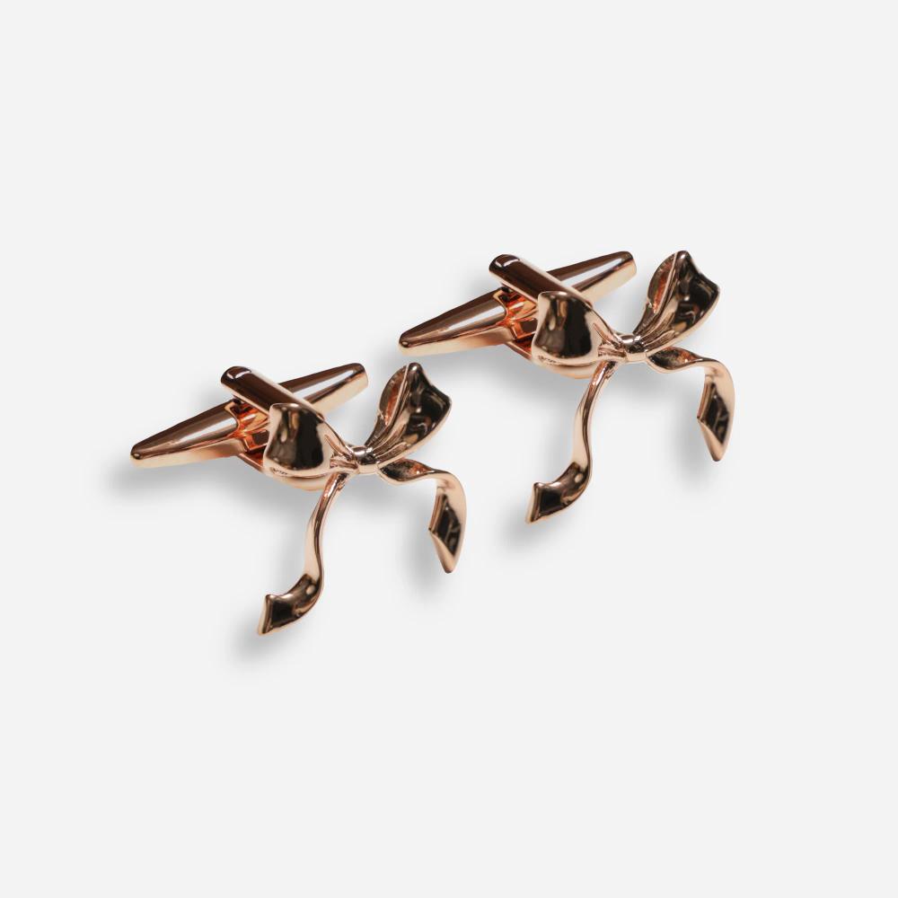 A pair of rose gold colored cuff links composed of a clip with a decorative bow on the front.