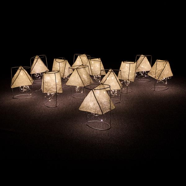 A dark image illuminated by a dozen small paper lanterns that sit on the floor. The lanterns are triangular in shape and are suspended from cylindrical armatures.