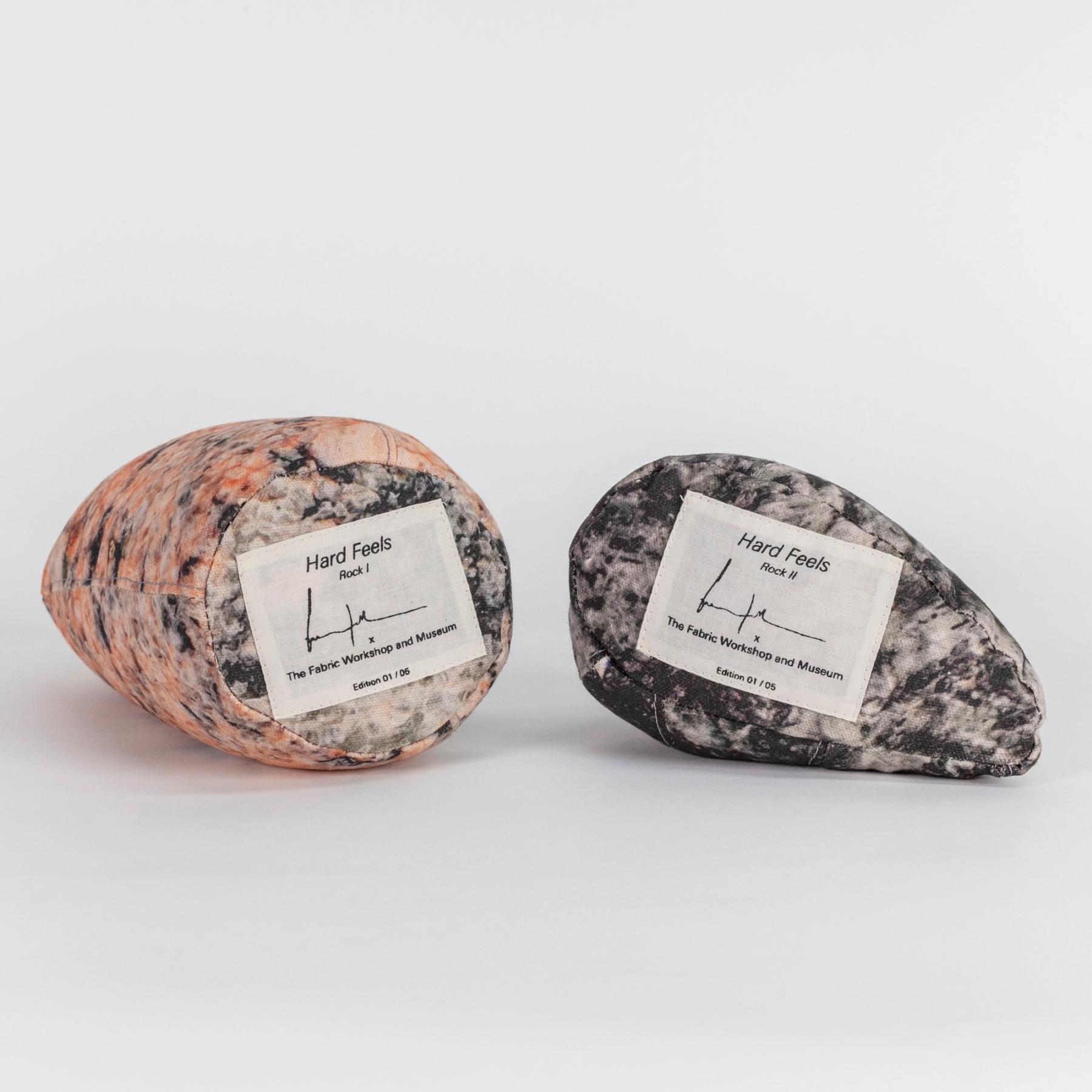 A photograph of the bottom sides of two small soft sculptures covered with patterns that resemble polished stone of different hues. Each one has a label reading the title of the edition (Hard Feels), the artist's signature, and edition number.