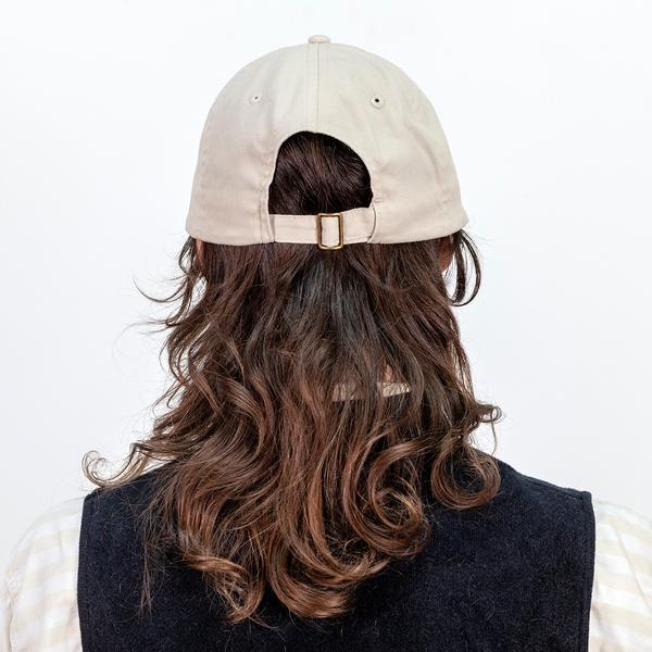 A photograph of a model turned away from the camera wearing a tan baseball cap with an adjustable strap.