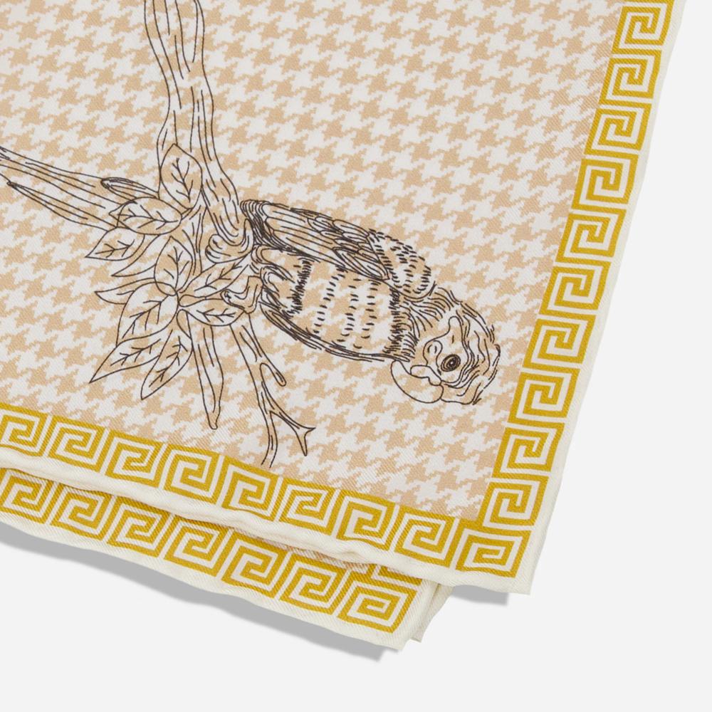 A folded pocket square with a detail of the corner. Its gold border resembles a Greek-style spiral frieze. There is a black line illustration of a Kasuku parrot sitting on a leafy branch on a light pink and white houndstooth background.