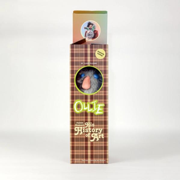 A photo of a tall brown plaid box with the top open, showing an image of a bearded man with dark bushy hair posing with a large gray rabbit. The box features a circular viewing window that reveals a gray stuffed animal inside. Above the viewing window it says "30 inch furry plushie" and includes a gold sticker that says "emotionally fulfilling!" Below the window it reads "Ollie" and "Jayson Musson's His History of Art".