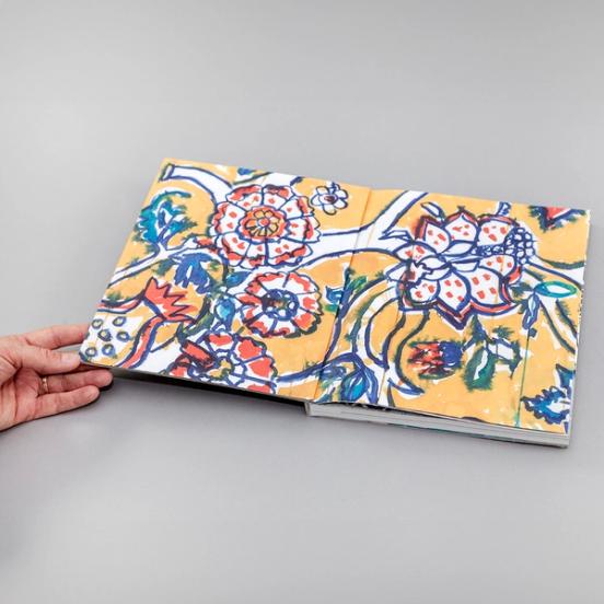 The image depicts a spread of a book. The book is open to a full-bleed two-page spread of a painterly drawing of bright flowers. There is a yellow background, and the flowers are outlined in blue and filled in with red, white, and orange.