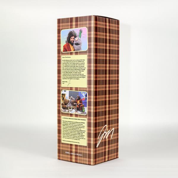 A photo of a tall brown plaid box in which two of its sides are visible. On the left are a couple images of a man with dark bushy hair wearing a burgundy coat and yellow turtleneck with a scraggily gray rabbit. Each image is accompanied with a yellow message. On the lower right of the box is the artist Jayson Musson's signature.