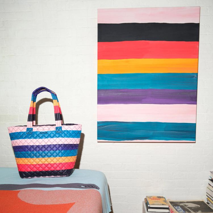 A photograph of a fabric quilted tote bag featuring painted horizontal stripes of alternating light pink, purple, blue, ochre, red, and black. The bag is sitting on a surface to the left of where a painting with the same horizontal striped pattern hanging on the wall.