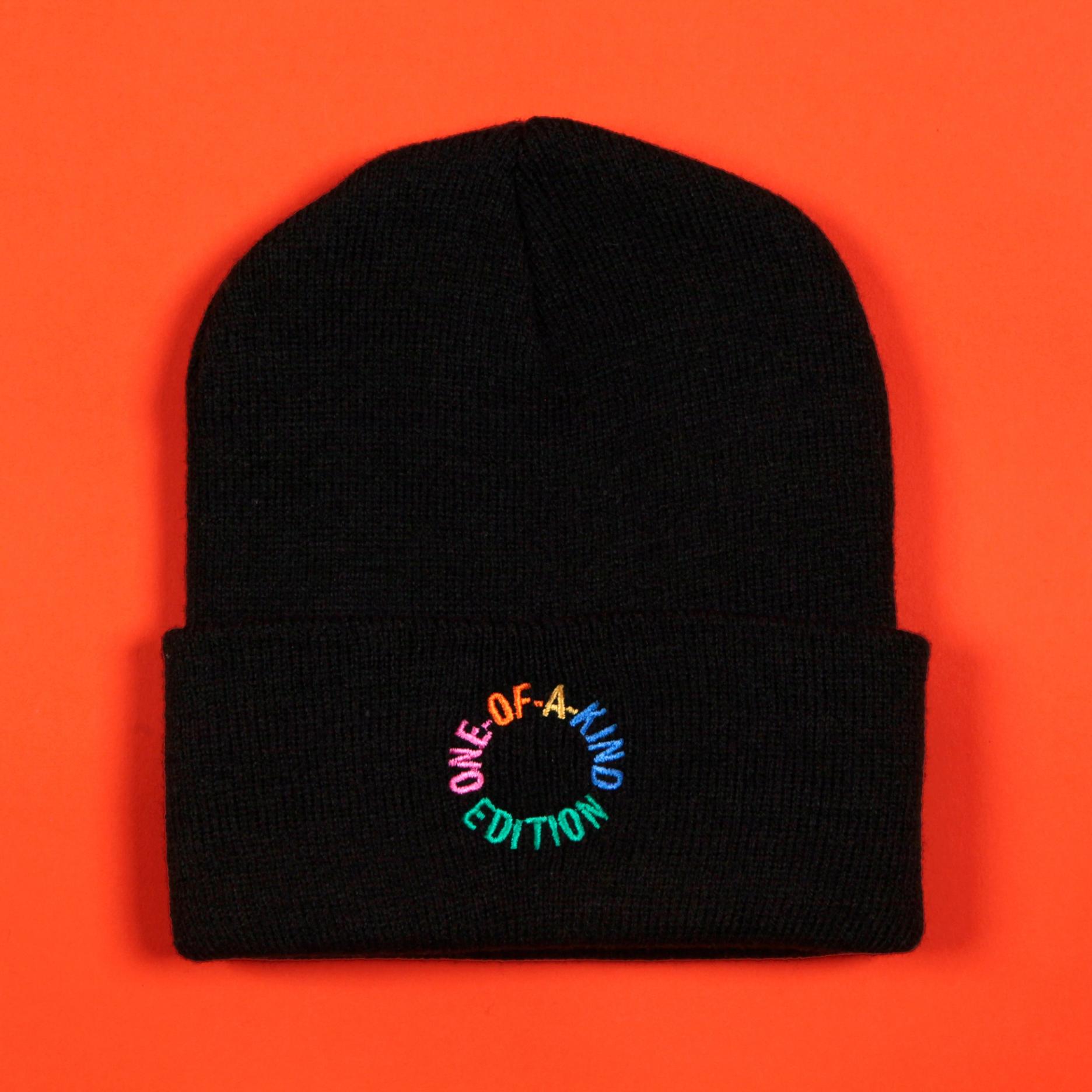 A photo of the black beanie against a tomato red background. The front of the beanie says "one-of-a-kind edition" in a circular text layout. Each word is a different color, in pink, orange, yellow, blue, and green. 
