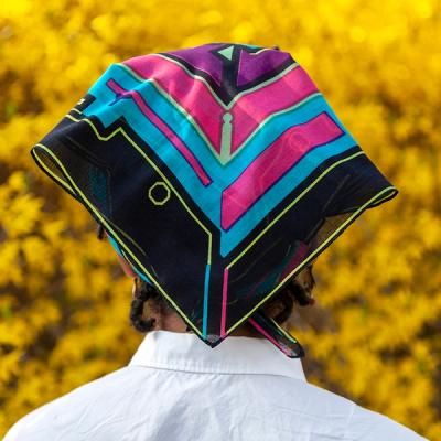 A person wearing a scarf on their head stands in front of yellow foliage, facing away from the viewer. This scarf is primarily black, blue, and bright pink, with a purple border around the inner design and neon green accents.