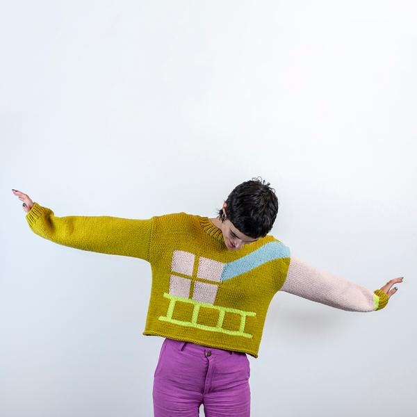 A person with short dark hair is wearing a long sleeve sweater with fuchsia-colored pants. She looks down as her hands spread outwards against a white background. The body of the sweater is a medium toned yellow green. The left sleeve is pink with a green cuff and a neon yellow accent next to the cuff. On the front of the sweater, there is the light pink implication of a window with an abstract beam of light in blue coming from the shoulder, and a neon yellow ladder lies flat across the bottom of the sweater, directly under the window.