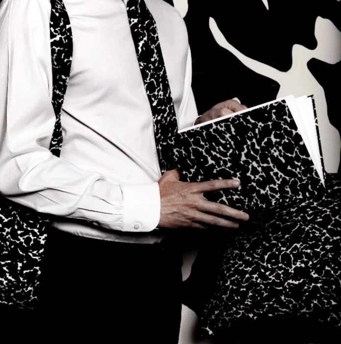A cropped image of a person dressed in a white dress shirt with a loose tie resting around their neck. The same speckled black and white design is featured on the tie, the lower background, and on a notebook the person is holding.