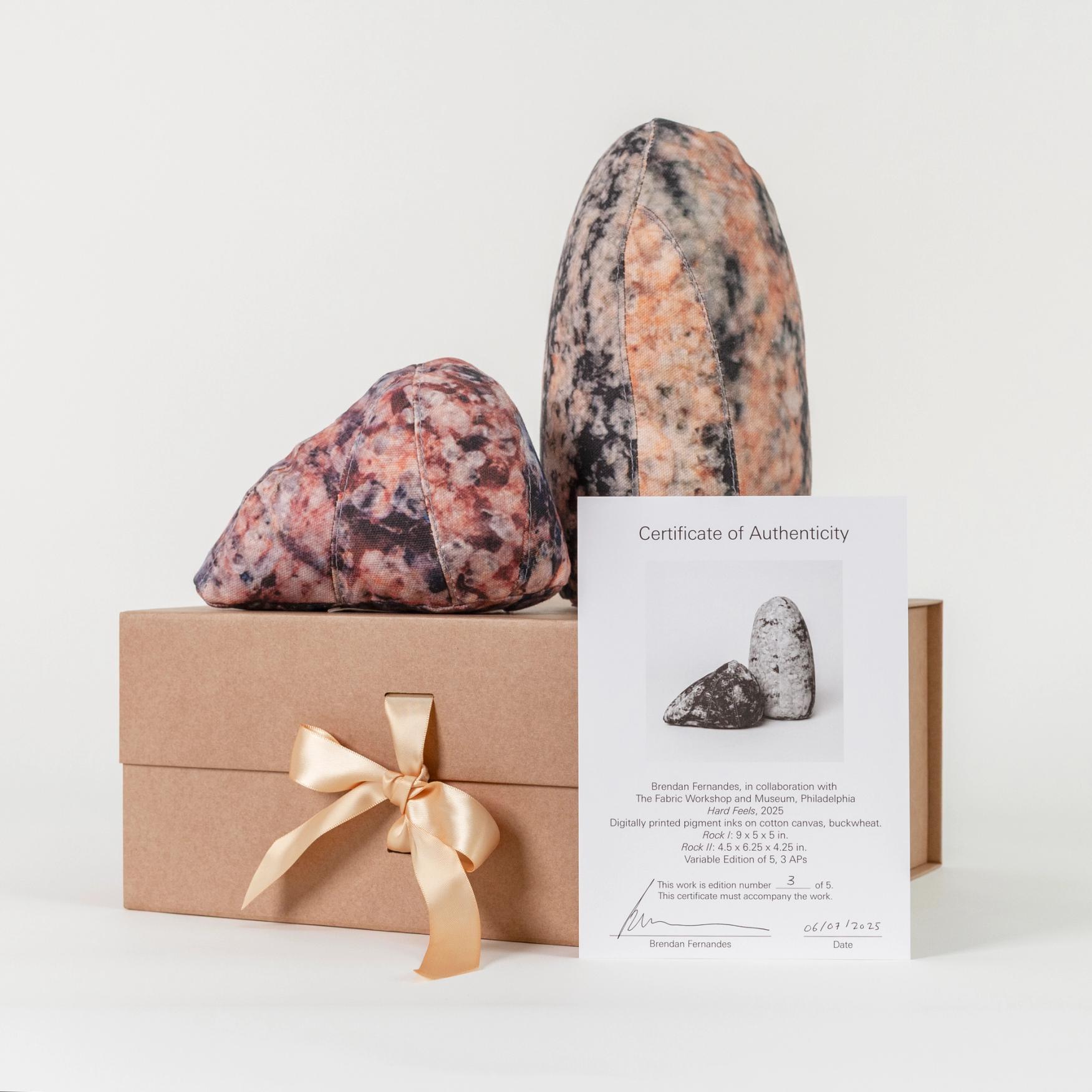 A photograph featuring two soft sculptures resembling polished stones, one short and triangular, the other tall and obelisk-shaped with a rounded top. The sculptures rest on a kraft paper gift box tied with a satin ribbon. In front of the box is a Certificate of Authenticity with an image of the sculptures and artist information.
