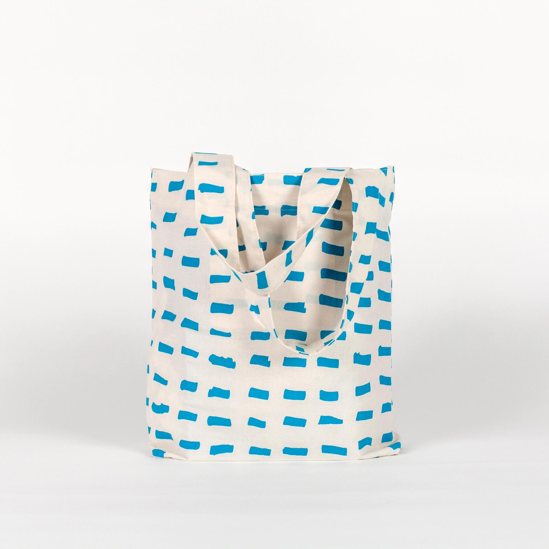 A tall, rectangular tote bag made of white fabric yardage with a printed pattern of short, gestural, brushstroke-like blue dashes.