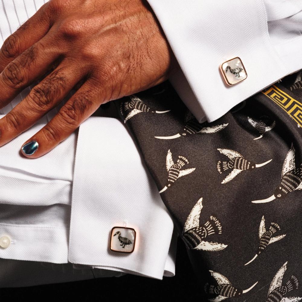 A detail of a person wearing a white dress shirt modeling the pair of square white pearlescent cufflinks with a rose gold border. On the pearlescent face is a black line illustration of a Bata bird.