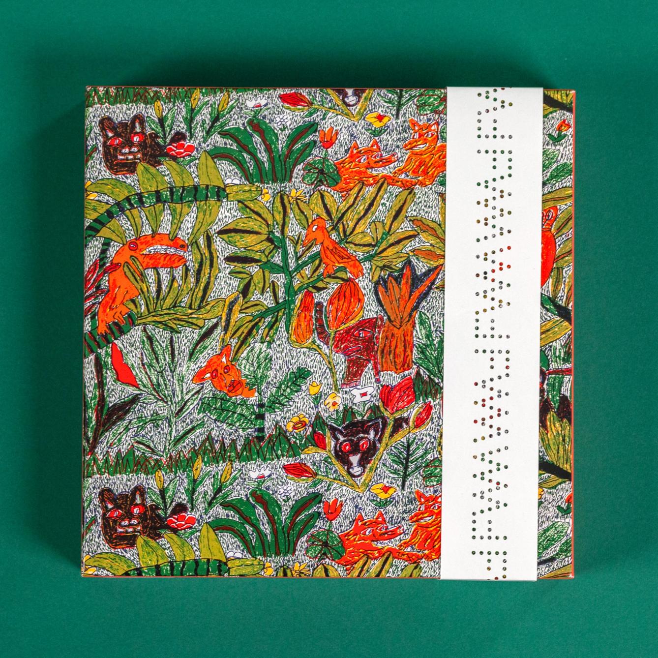 A puzzle box is on top of a dark green background. The puzzle box shows the pattern of the puzzle, which is a hand-drawn repeat design featuring illustrative jungle plants and animals in vibrant oranges and greens. On the right is a white paper band of packaging with the FWM logo cut out in a repeating strip.